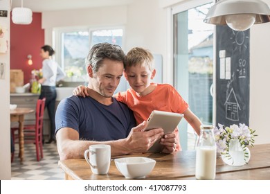 Papa und sein 8-jähriger Sohn benutzen beim Frühstück in der Küche eine digitale Tablette, Mutter arbeitet auf unscharfem Hintergrund.  Sie tragen ungezwungene Kleidung