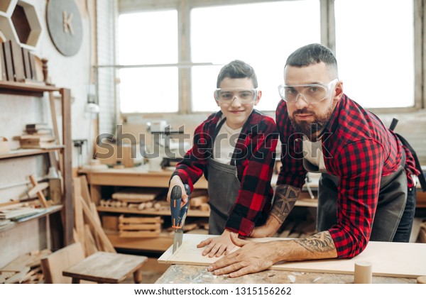 Dad and his\
son dressed in checkered red shirts and wearing safety glasses are\
working in the carpentry workshop, son is learning to work with a\
saw under father s\
supervision.