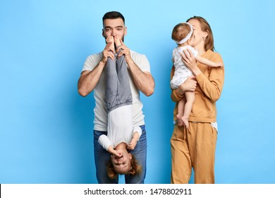 Dad Holding His Child Upside Down And Kissing Her Feet, While His Wife Kissing Baby On Cheek. Isolated Blue Background. Studio Shot