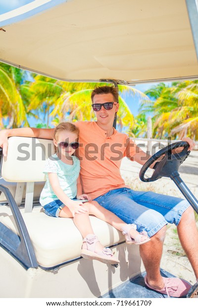 Dad and little daughter driving golf cart on\
tropical beach