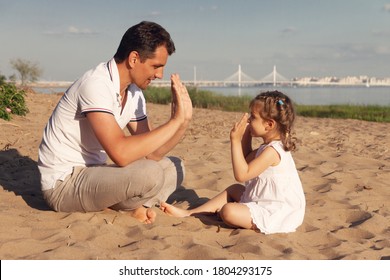 Dad And Little Daughter In White Dress Are Sitting On Beach And Playing With Their Palms On Sunny Day