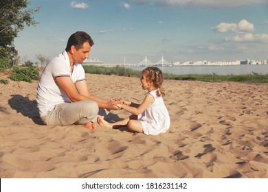 Dad And Little Daughter In White Dress Are Sitting On Beach Of Baltic Sea And Playing With Their Palms, Copy Space