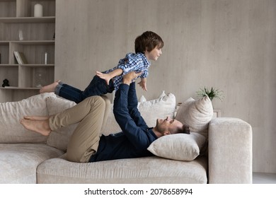Dad lying on couch lifts on outstretched hands little 6s son. Happy cute boy play plane fly in air while his father raise him up on arms. Active weekend at home, dreams about holiday, leisure concept