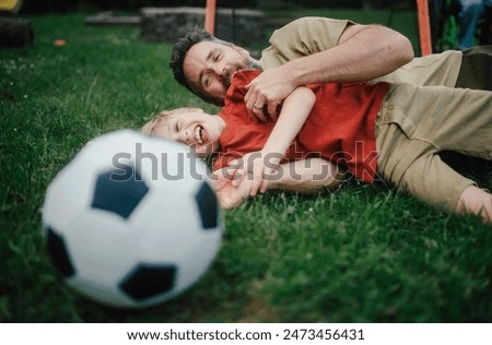 Dad lying on grass with son, laughing and having fun. Father and boy playing football, catching each other, falling to the ground. Fatherhood and Father's Day.