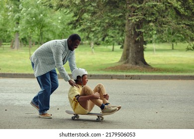 Dad Pushing His Son In Back And Rolling Him While He Sitting On Skateboard, They Riding Along The Road In Park