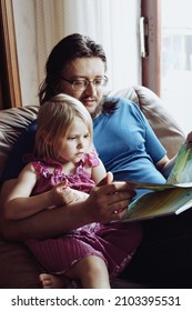 Dad Reading Bedtime Story To His Little Daughter. They Are Sitting On Big Comfy Armchair Near Window