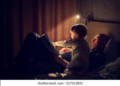 Dad is reading a bedtime story to his little son. Night lamp is shine beautifully. Image with selective focus and toning
