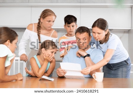 Dad is sitting in kitchen, reading letter and is upset by unpleasant and news. Wife, daughters and children are worried, look carefully at ass and wait for explanation