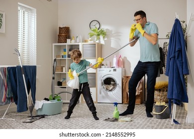 Dad and son dance in the laundry room, playing cleaning accessories. The little boy holds the mop in hands like a guitar, the man plays the floor brush like a harmonica, housekeeping.