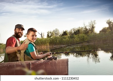 Dad And Son Fishing From Wooden Boat On River, Happy Family Together In Nature