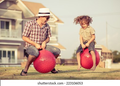 Dad And Son Jumping On Inflatable Balls On The Lawn In Front Of House At The Day Time