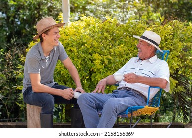 Dad and son laughing and drinking coffee on a sunny morning. Latin American peasants in work clothes having a relaxing time. Elderly man with his young son.