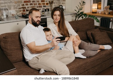 A Dad, A Son, And A Mom Are Watching A Video On A Smartphone On The Sofa. A Father With A Beard Is Demonstrating To His Family News On The Screen Of The Cell Phone In The Evening At Cozy Home.