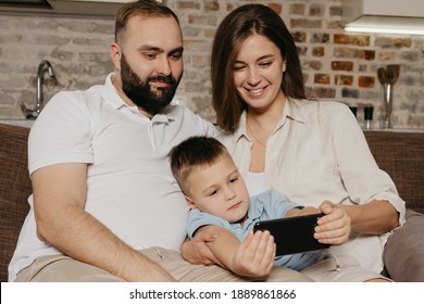 A dad, a son, and a mom are watching video on the sofa. A child is demonstrating the show on the smartphone to smiling mother and father in the evening at home. - Shutterstock ID 1889861866