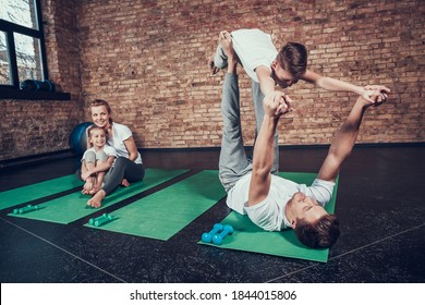 Dad And Son Perform Acrobatic Exercise In The Gym. A Man Lies On The Mat And Holds His Son While Mom And Daughter Look And Smile. 
