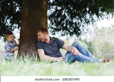 Dad And Son Play Hide And Seek Sitting Under The Tree In The Park