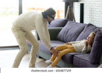 Dad and son playing hide and seek game at home, blindfolded father trying to catch excited happy child boy hiding on couch, parent and kid having fun laughing spending time together in living room