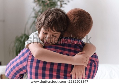 Dad and son sharing a joyful hug, seated on a sofa in the living room, grinning towards the lens. The Significance of Family Connection and Cherished Moments. Father and Son Bonds
