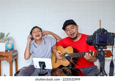 Dad and son singing and playing guitar together while doing video online streaming. Asian family spending quality time at home while recording video on digital camera.