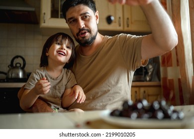 Dad And Son Sit In The Kitchen At Home And Eat Cherries. The Concept Of A Happy Family. Shallow Depth Of Field