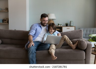 Dad And Son Sit On Sofa Enjoy Weekend Use Laptop, Rest At Home Spend Leisure On Internet, Buying Goods, Make Order On-line, Choose Cartoons Or Family Movies On Digital Streaming Services. Tech 