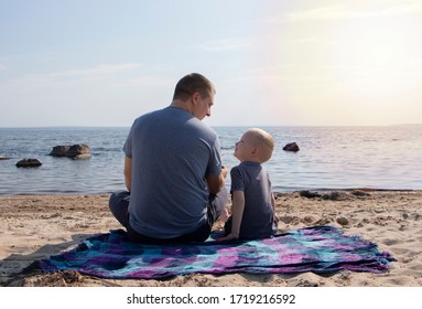 Dad And Son Are Sitting On The Beach And Chatting. A Boy With A Father On The Background Of The Sea And Sunset Cuddling. The Concept Of A Happy Childhood And Hanging Out With A Parent