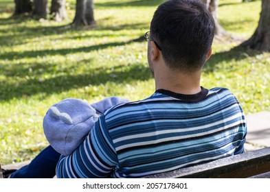 Dad In A Striped Pullover Is Holding A Newborn In Arms, On A Park Bench. The Baby Is Sleeping And Wears A Bear Form Costume.nature Background, Green Grass, Space For Text, Phone