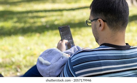 Dad In A Striped Pullover Is Holding A Newborn In Arms, On A Park Bench. The Baby Is Sleeping And Wears A Bear Form Costume.nature Background, Green Grass, Space For Text, Phone