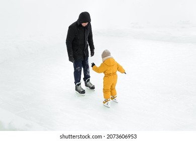 Dad teaches daughter to skate on the frozen ice of the lake, standing in front of the child. Father and the girl have fun at the ice rink on a snowy frosty winter day. Support and support in endeavors