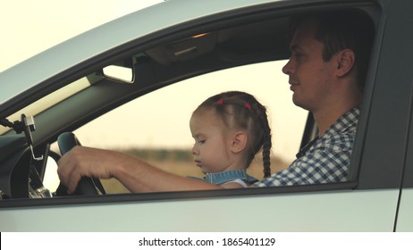 Dad Teaches His Little Daughter To Turn Steering Wheel While Sitting In His Car In Drivers Seat. Father Travels With Children By Car. Driver And Kid Are Driving. Happy Family And Childhood Concept.