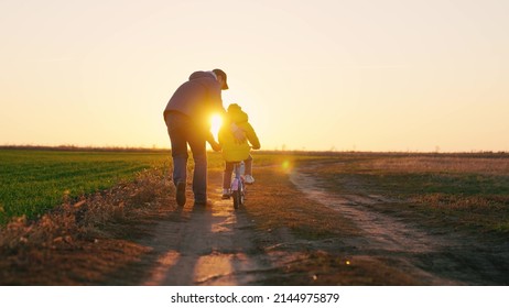 Dad teaches a little girl to ride a children's bike on the road in spring. Happy family. A father teaches his daughter to ride a bike in the park at sunset. Daddy and kid play together outdoors
