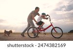 dad teaches son to ride a bike. happy family kid dream concept. the boy sat on bicycle for the first time, his father teaches boy to ride a bicycle. dog runs with family, fun family pastime lifestyle