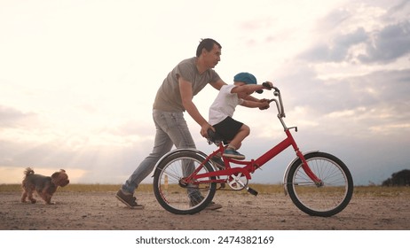 dad teaches son to ride a bike. happy family kid dream concept. the boy sat on bicycle for the first time, his father teaches boy to ride a bicycle. dog runs with family, fun family pastime lifestyle - Powered by Shutterstock