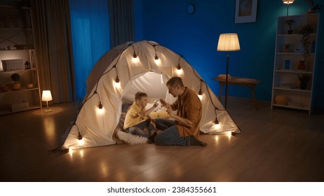 A dad is teaching his son to count. Man shows fingers on hands, that boy is counting. Father sits on floor near tent in living room where boy is sitting. Concept of parenting, teaching.