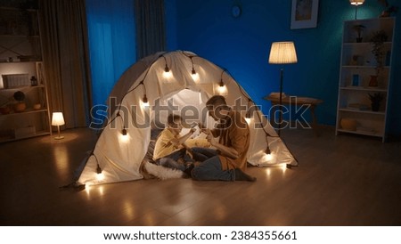A dad is teaching his son to count. Man shows fingers on hands, that boy is counting. Father sits on floor near tent in living room where boy is sitting. Concept of parenting, teaching.