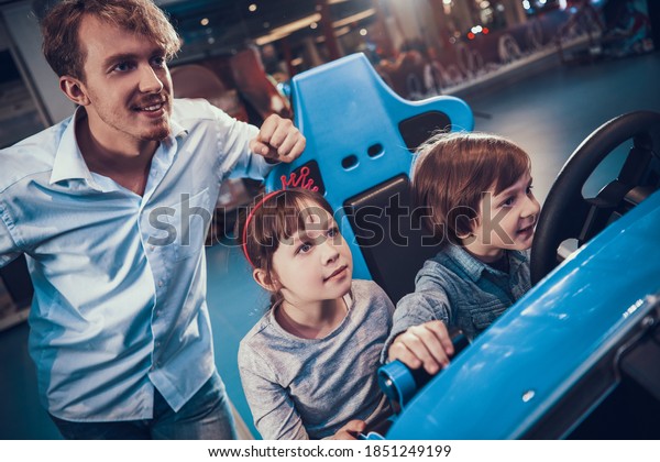 Dad watches his daughter and son drive a toy car\
in a video game. Children sit together in the entertainment center\
and play a video game. 