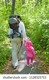 A Dad Wearing An Empty Baby Carrier On His Back Walks On A Forest Path Sweetly Holding Hands With A Toddler Girl Dressed In Pink. Photo Taken In Wisconsin.