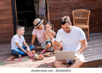 Dad Works On Family Vacation, Uses A Laptop Sitting On The Porch Of A Wooden Country House, In The Background His Wife And Two Children