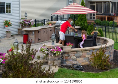Daddy serving lemonade to his little daughter outdoors on a brick patio as she relaxes in the shade of an umbrella near an outdoor kitchen and gas barbecue in summer