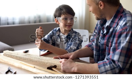 Daddy teaching his smiling son carpentry, little boy using screwer at workshop