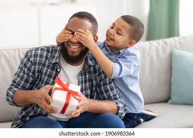 Dad's Birthday. Little Son Congratulating Black Father Giving Wrapped Gift To Him, Covering Eyes Sitting On Couch At Home. Family Holiday Celebration, Surprise Present For Daddy's B-Day - Powered by Shutterstock