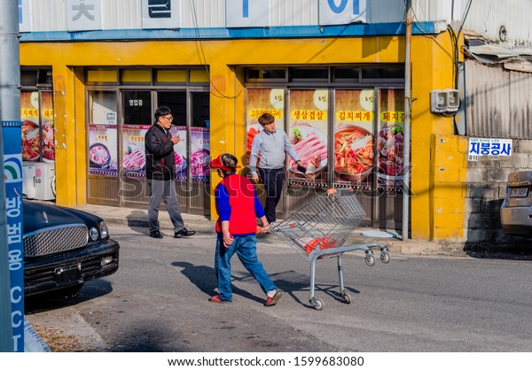 Daejeon,\
South Korea; December 24, 2019: Unidentified woman wearing red vest\
pulling shopping cart down paved street in front of two\
unidentified men walking in front of\
restaurant.