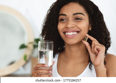 Daily Intake Of Vitamins And Minerals. Happy Black Woman Holding Capsule With Beauty Supplements And Water Glass, Closeup