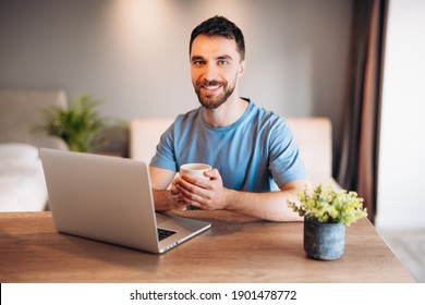 Daily life of young man in kitchen using laptop holding a cup of coffee. Attractive male working from home, telecommuting using remote internet job online communication on. - Powered by Shutterstock