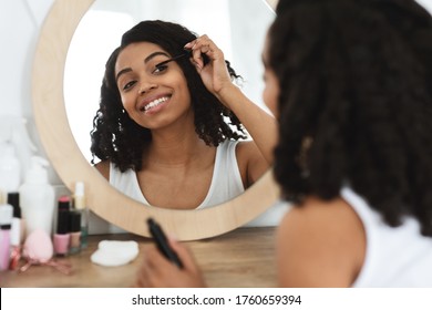 Daily Makeup. Beautiful Black Woman Applying Mascara On Eyelashes At Home, Looking In The Mirror On Dressing Table, Over Shoulder View