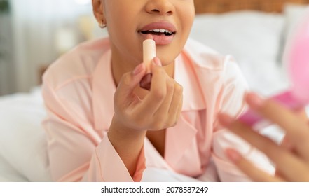 Daily Makeup Routine. Unrecognizable Black Lady In Pajamas Applying Lipstick Holding Tube, Getting Ready For Work, Closeup. Woman Lying On Bed In Bedroom, Using Natural Decorative Cosmetics