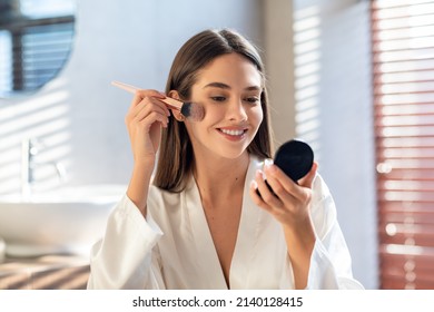 Daily Makeup. Smiling Young Female Applying Blush With Makeup Brush In Bathroom, Beautiful Happy Woman Wearing White Silk Robe Using New Cosmetics While Getting Ready At Home, Copy Space