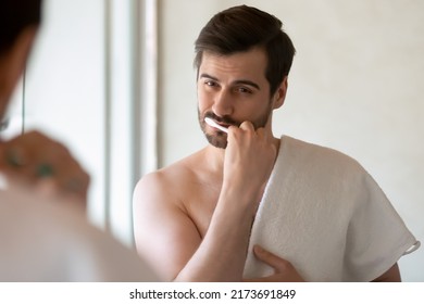 Daily routine. Concentrated handsome young man standing shirtless at bathroom holding towel on shoulder looking at mirror brushing cleaning teeth having healthy habit to begin day from dental hygiene Foto Stock