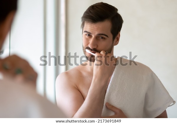 Daily routine. Concentrated handsome young man\
standing shirtless at bathroom holding towel on shoulder looking at\
mirror brushing cleaning teeth having healthy habit to begin day\
from dental hygiene