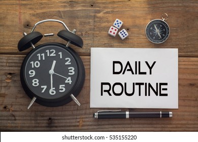 Daily Routine written on paper with wooden background desk,clock,dice,compass and pen.Top view conceptual